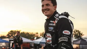 Isaac Johnson in a black-and-white firesuit smiles at the track at sunset with tents, equipment, and another person blurred in the background.