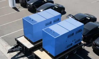 Two blue large box mobile chargers parked along a row of black Tesla EV sedans in a parking lot.