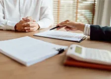 Two people sit across from each other at a desk during a business meeting. One person, wearing a white shirt, has their hands folded while the other gestures with a pen toward documents clipped to a clipboard. Additional paperwork and a calculator are visible on the table, suggesting a discussion involving contracts, finances, or administrative paperwork. Sunlight filters through window blinds in the background, creating a professional office setting.