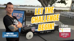 Lauren Fletcher stands with arms crossed beside a black soap box derby car at a waterfront venue. Text on image reads “Let the Challenge Begin” Work Truck, Hourglass Foundation, and Soap Box Derby