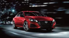 Red 2027 Nissan Altima SR sedan driving through a city at night, shown from the front three-quarter angle with illuminated headlights and dark alloy wheels.