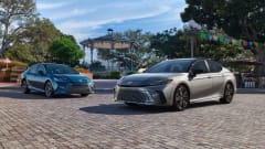 Two 2027 Toyota Camry sedans, one silver and one blue, parked on a cobblestone plaza with a gazebo, trees, and colorful decorations in the background.