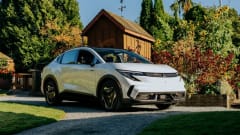 White 2027 Mitsubishi Eclipse Sportback shown from the front and side on a gravel driveway beside a garden and wooden shed.