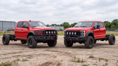 Red Ram and Ford crew cab chassis trucks parked on a gravel lot, with oversized off-road tires, front winches, auxiliary lights, and exposed rear frame rails.