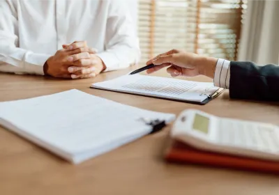 Two people sit across from each other at a desk during a business meeting. One person, wearing a white shirt, has their hands folded while the other gestures with a pen toward documents clipped to a clipboard. Additional paperwork and a calculator are visible on the table, suggesting a discussion involving contracts, finances, or administrative paperwork. Sunlight filters through window blinds in the background, creating a professional office setting.