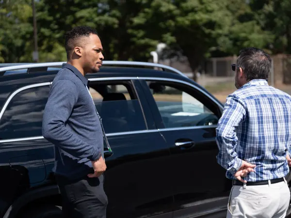 Two men converse next to a vehicle in ride and drive. 