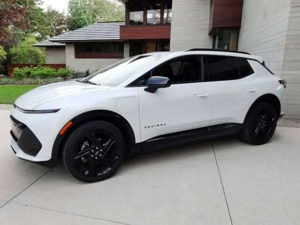 A white Chevrolet Equinox EV, small SUV, parked on the driveway outside a large, modern looking house.