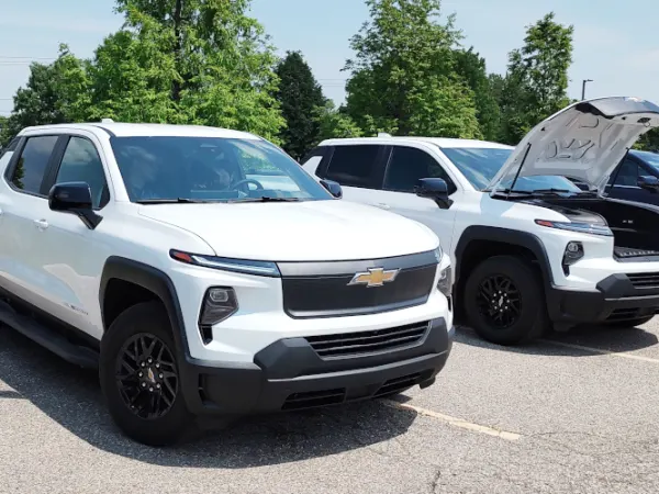 Two white EV pickups are parked, the one further from the camera has the front hood open.
