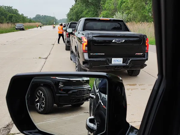 View out driver's window looking over the side-view mirror at other black pickup trucks waiting in line ahead.