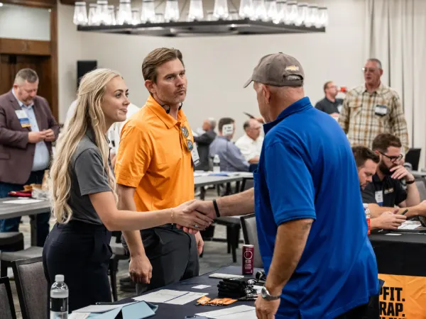 A woman in a gray shirt shakes hands with a man in a blue polo across a table at Work Truck Exchange, while another attendee looks on.