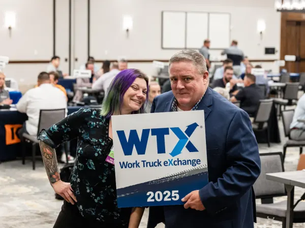 Lauren Fletcher and Wayne Parham posing with an attendee holding a Work Truck Exchange 2025 sign inside the meeting room filled with one-on-one business meetings.