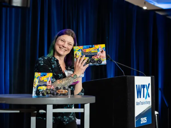 Lauren Fletcher smiling on stage while holding Hot Wheels prize boxes during the Work Truck Exchange 2025 Trivia Luncheon.