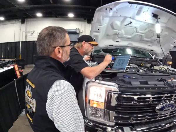 One man watching another work under the hood of a large pickup truck.