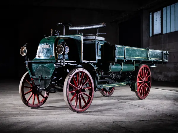 1899 Daimler truck with red spoked wheels and early shaft-drive system displayed in a dark studio setting.