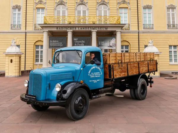 Blue vintage Mercedes-Benz flatbed truck parked in front of historic museum building, representing mid-century commercial vehicles.
