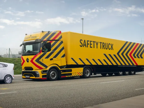 Yellow Mercedes-Benz eActros 600 Safety Truck with bold safety graphics driving on a test track beside a passenger vehicle.