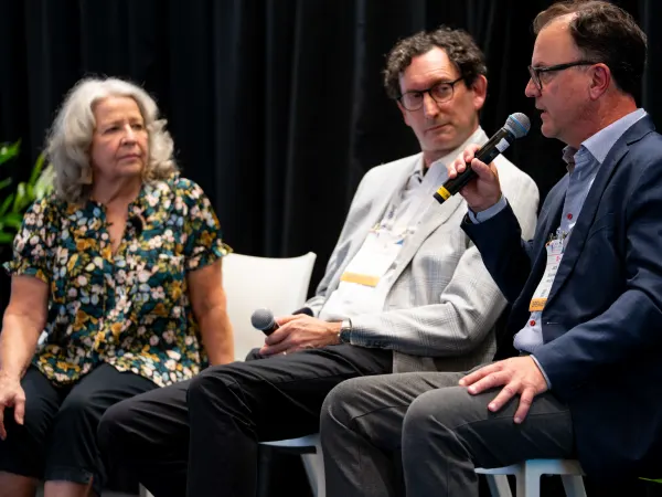 Three panelists participate in a discussion at the 2026 Fleet Forward Conference, with one speaker addressing the audience using a handheld microphone while the other two listen.