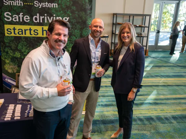 Three conference attendees pose for a photo in front of a Smith System safe driving display at a fleet industry event.