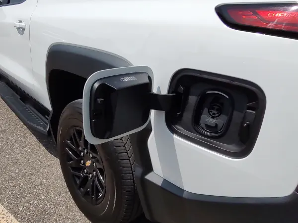 View of the left rear corner of a white EV truck with the door covering charging ports open.