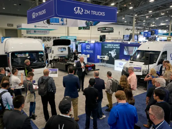 Media gather on show floor to listen to a press conference, trucks in background. 