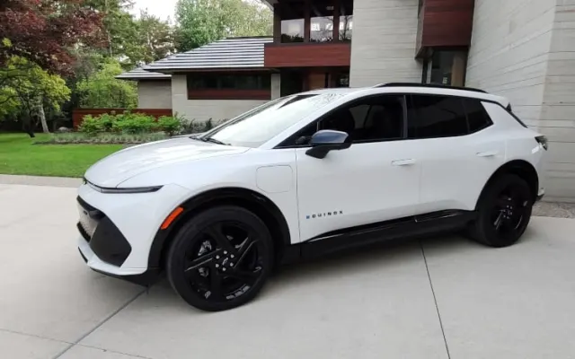 A white Chevrolet Equinox EV, small SUV, parked on the driveway outside a large, modern looking house.