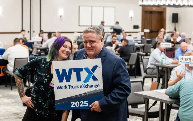 Lauren Fletcher and Wayne Parham posing with an attendee holding a Work Truck Exchange 2025 sign inside the meeting room filled with one-on-one business meetings.
