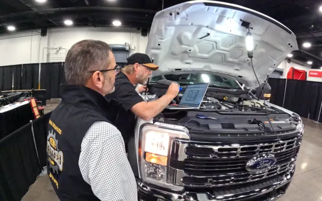 One man watching another work under the hood of a large pickup truck.
