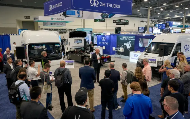 Media gather on show floor to listen to a press conference, trucks in background.