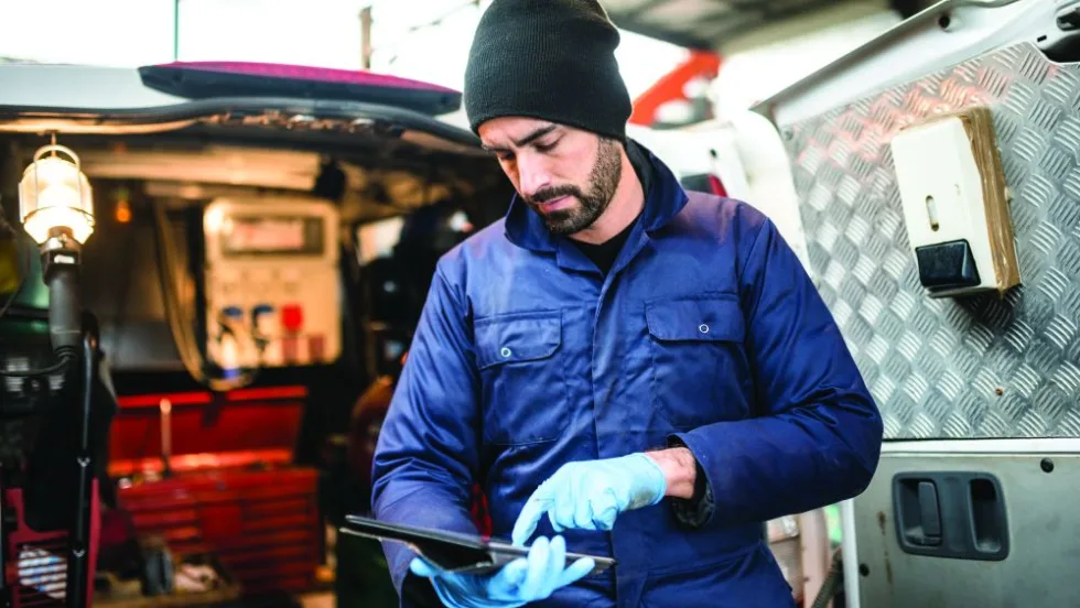  Fleet technician wearing gloves and a blue work jacket reviews information on a tablet inside a mobile service vehicle.