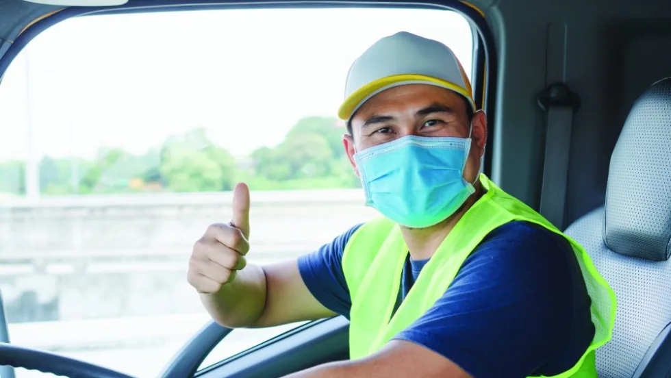  Delivery driver wearing a face mask and high-visibility vest gives a thumbs-up from the cab of a truck.