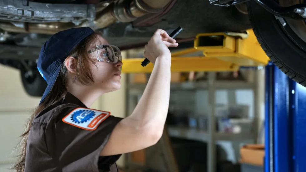Female automotive mechanic uses a flashlight to check under a vehicle overhead on a lift.