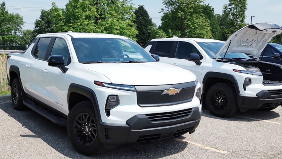 Two white EV pickups are parked, the one further from the camera has the front hood open.