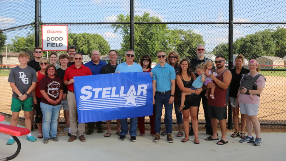 Group photo in front of backstop of a baseball field as people hold a banner that says Stellar.