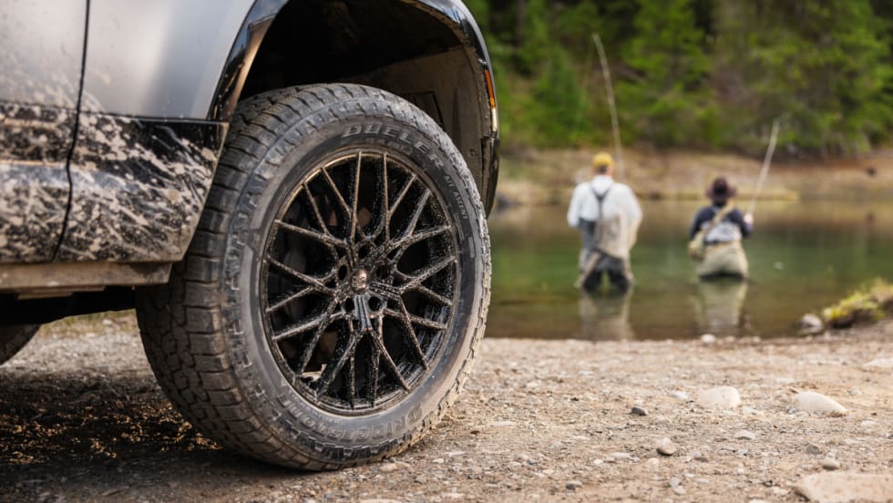 An SUV's tire is showcased beside a calm body of water, highlighting the vehicle's rugged design in a natural setting.