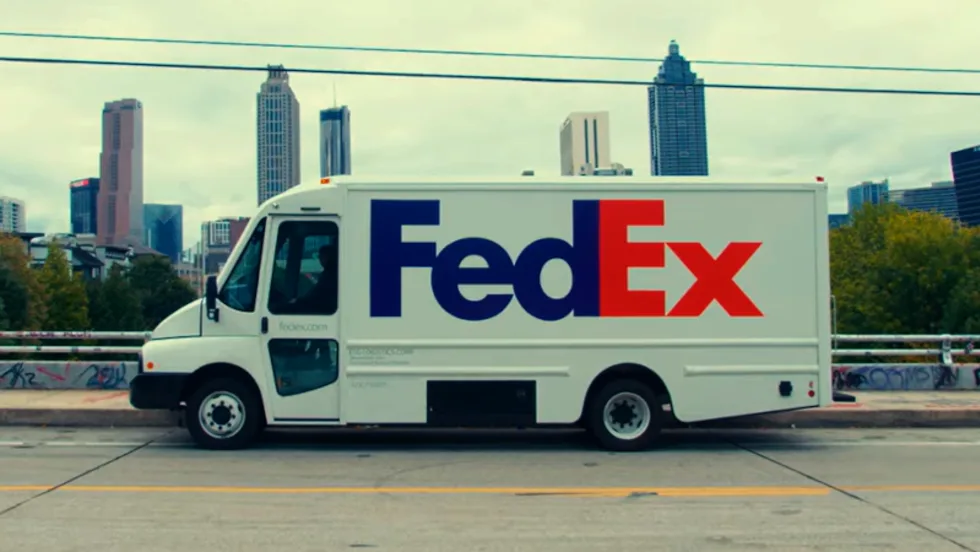 White battery-electric FedEx truck parked in front of a city skyline.