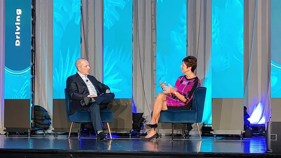 man and woman in business attire have a seated conversation on stage.