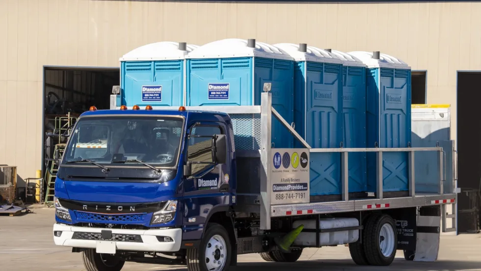 Battery electric truck with blue cab and flatbed hauling portable toilets.