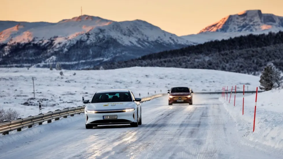 A Lucid Air Grand Touring drives on a snow-covered road in Norway during the NAF Winter Test, with mountains in the background and winter conditions visible.