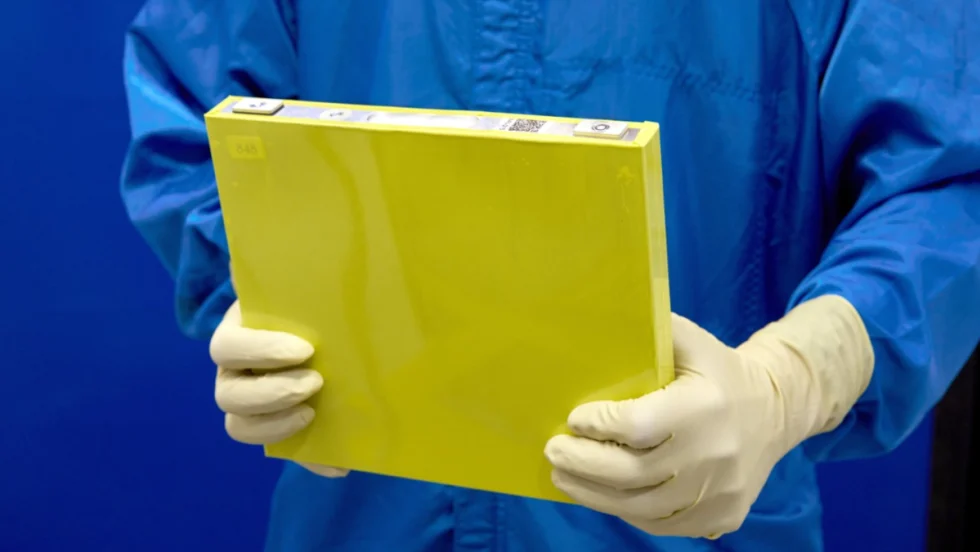 Closeup of person in blue lab clothing wearing gloves and holding a yellow battery cell.