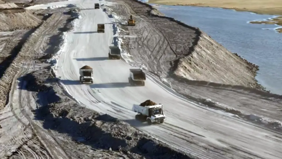 View of mining dump trucks driving on a curved road on top of a hill next to water.