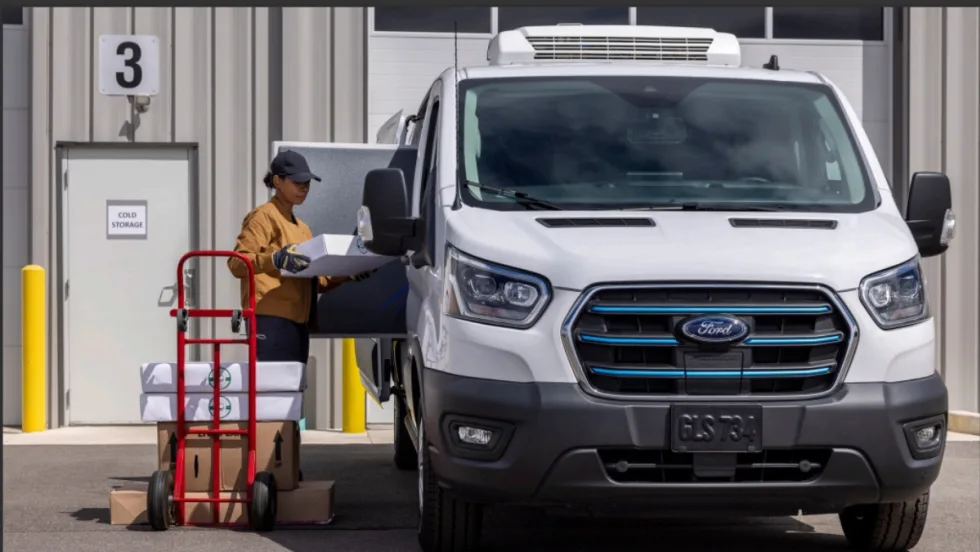 A delivery worker unloads boxes from a Ford electric refrigerated van parked outside a cold storage facility, illustrating electric truck use in commercial fleet operations.