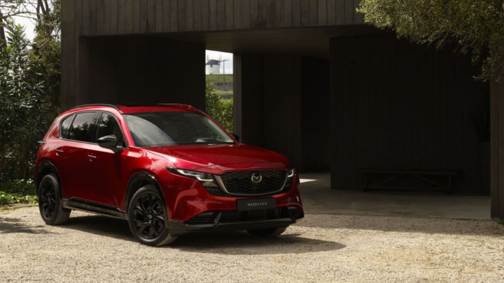 Front three-quarter view of a red 2026 Mazda CX-5 parked on a gravel driveway in front of a modern, dark wood building, featuring black wheels and Mazda's Signature Wing grille.