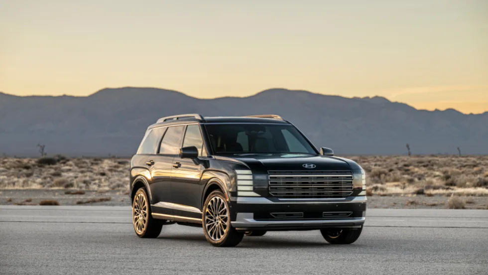 Front three-quarter view of the 2026 Hyundai Palisade SUV parked on a desert road at sunset, with mountains in the background.
