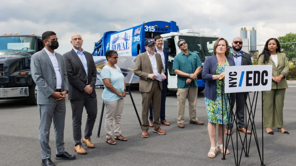 Group of officials standing around woman making announcement with two battery-electric trucks in the background.