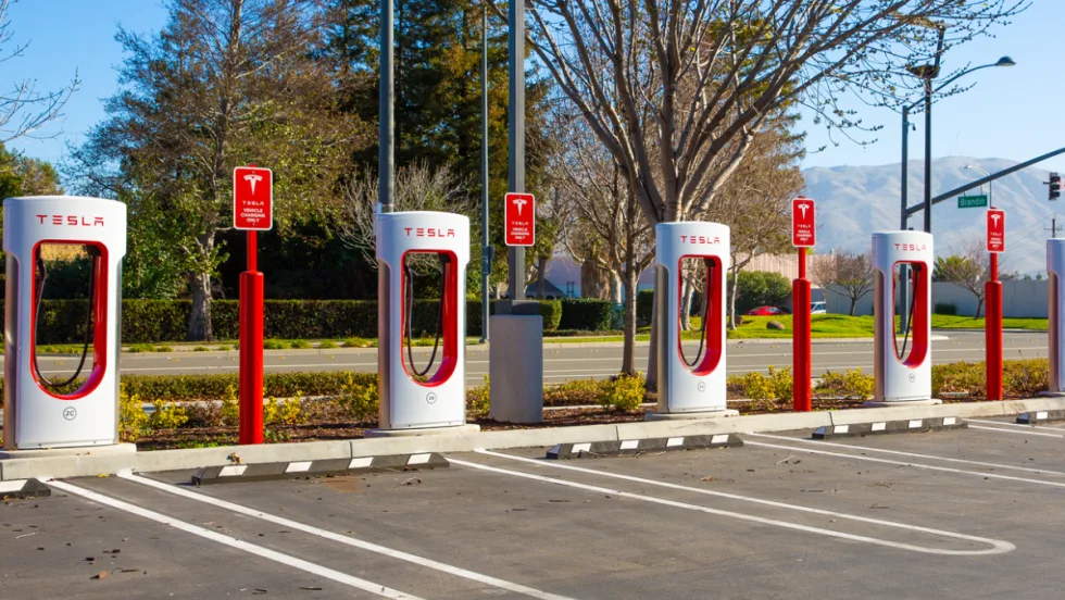 Row of Tesla Supercharger stations in a parking lot, ready for electric vehicles to charge, with clear skies and mountains visible in the background.