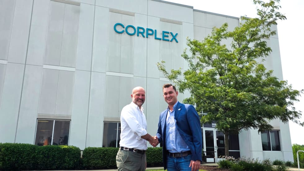 Two businessmen shake hands outside an industrial building with a Corplex name on the upper part of the building.