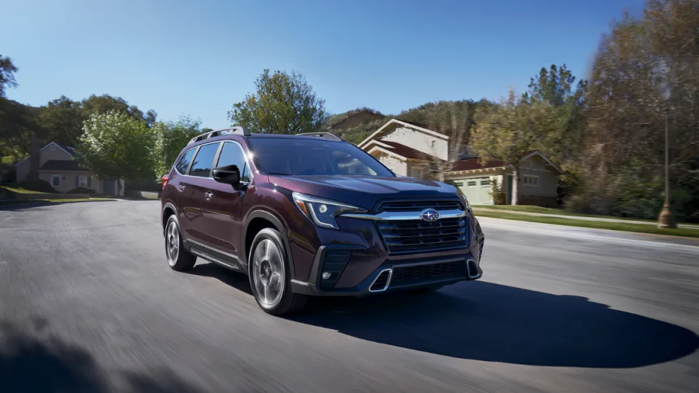 A maroon 2026 Subaru Ascent three-row SUV with a bold black front grille, LED headlights, and roof rails drives through a suburban neighborhood on a sunny day.