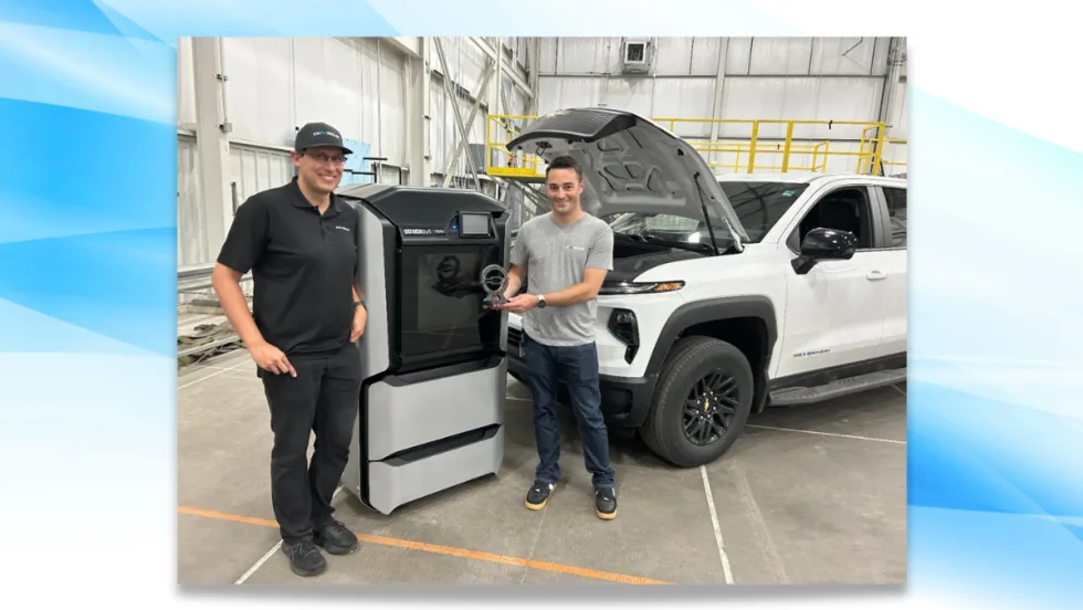 Two GM engineers stand beside a 3D printer holding a trophy, with a white Chevrolet Silverado EV Max Range Work Truck in the background and hood open after a record-setting range test.