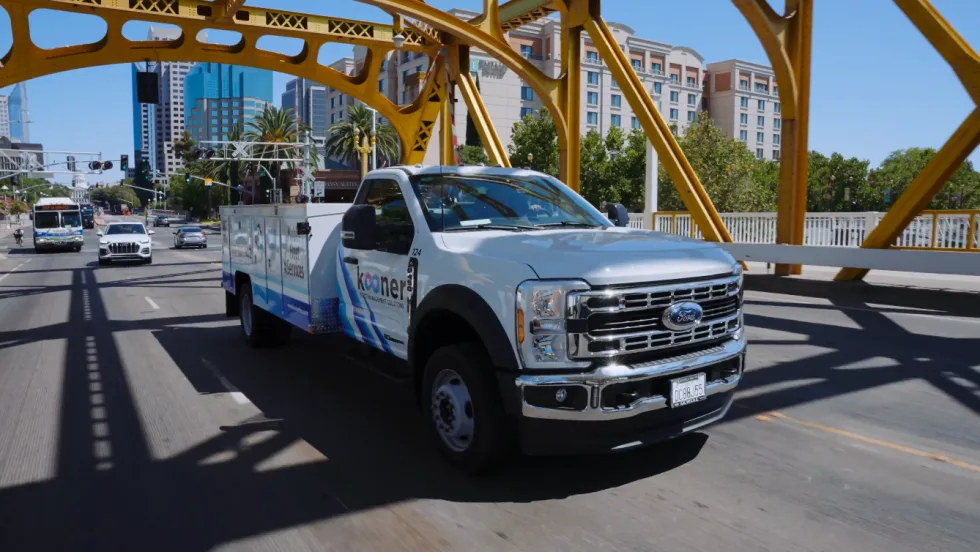 A white service truck driving across a yellow bridge with city skyline in the background.