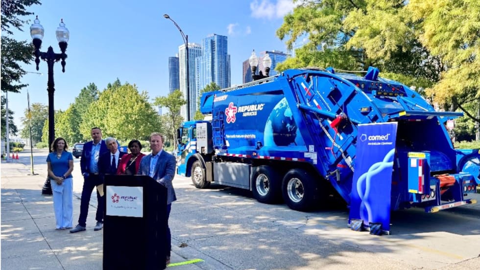Group of officials during an event showcasing a blue Mack LR Electric refuse truck, one speaker is at the podium talking and others are in the background. 
